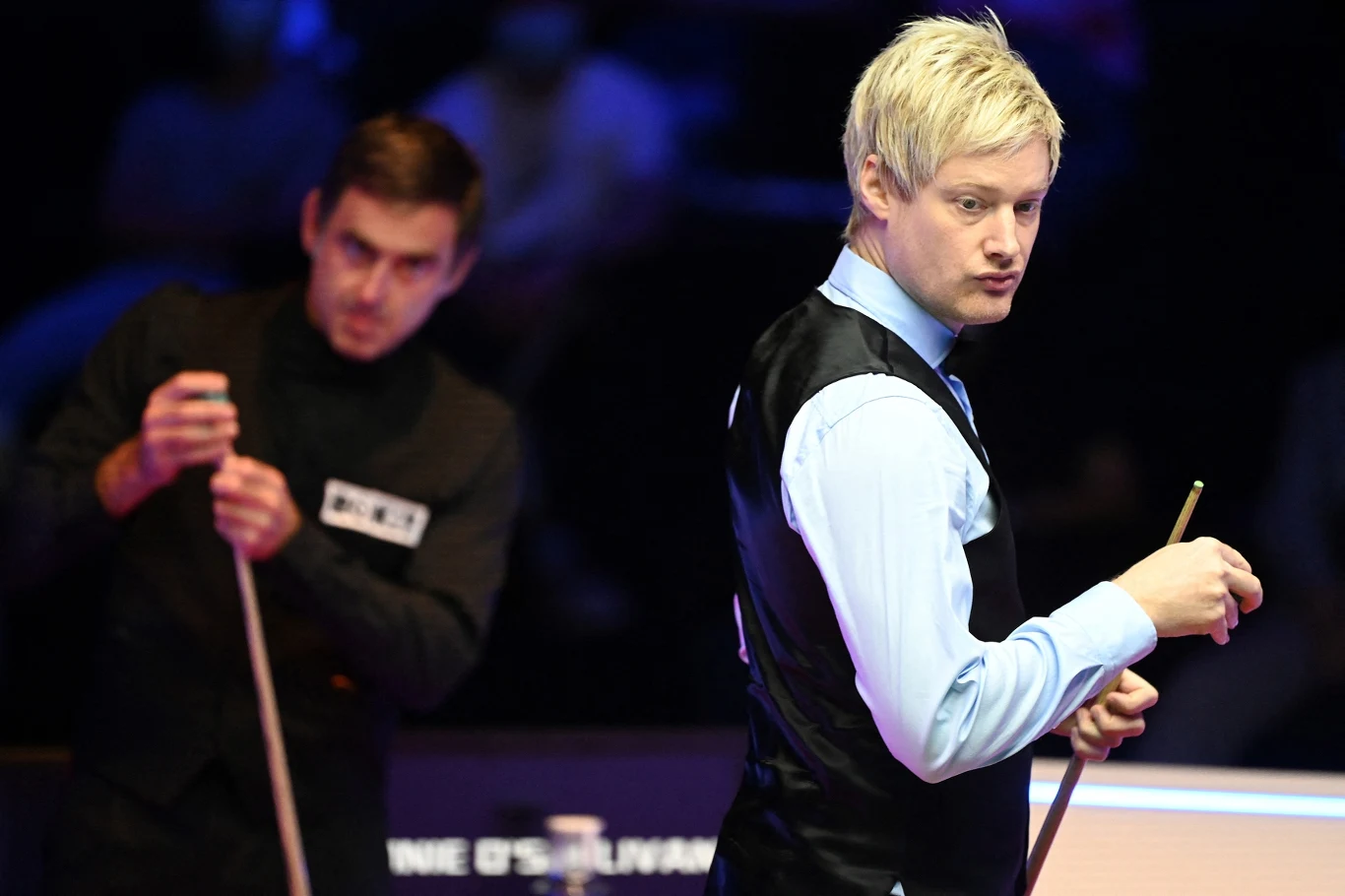 Dwóch zawodników snookera przy stole bilardowym, jeden z nich koncentruje się na swoim ruchu, drugi w tle obserwuje sytuację. W tle zamazana publiczność tworzy sportową atmosferę.