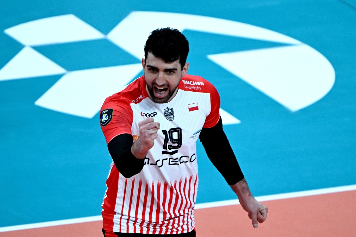 Zawodnik siatkarski w biało-czerwonym stroju z numerem 19 na koszulce, energicznie zaciska pięść w geście triumfu na tle niebieskiego boiska sportowego.