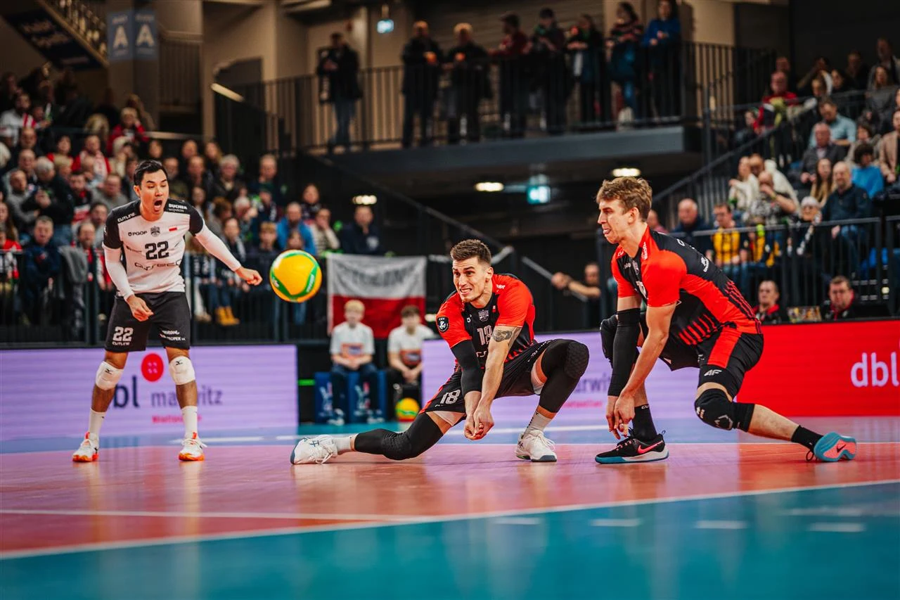 Trzech siatkarzy podczas meczu halowego, dwóch z nich w czerwono-czarnych strojach kuca i koncentruje się na odbiorze piłki, tłum kibiców w tle oraz kolorowe bandy reklamowe przy boisku.