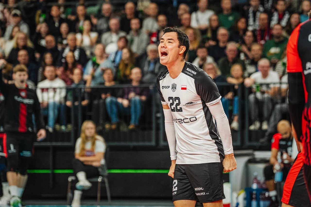 Zawodnik siatkarski w czarno-białym stroju z numerem 22 wyraża silne emocje na tle pełnej widowni podczas meczu, wokół znajdują się inni gracze i osoby związane z wydarzeniem sportowym.