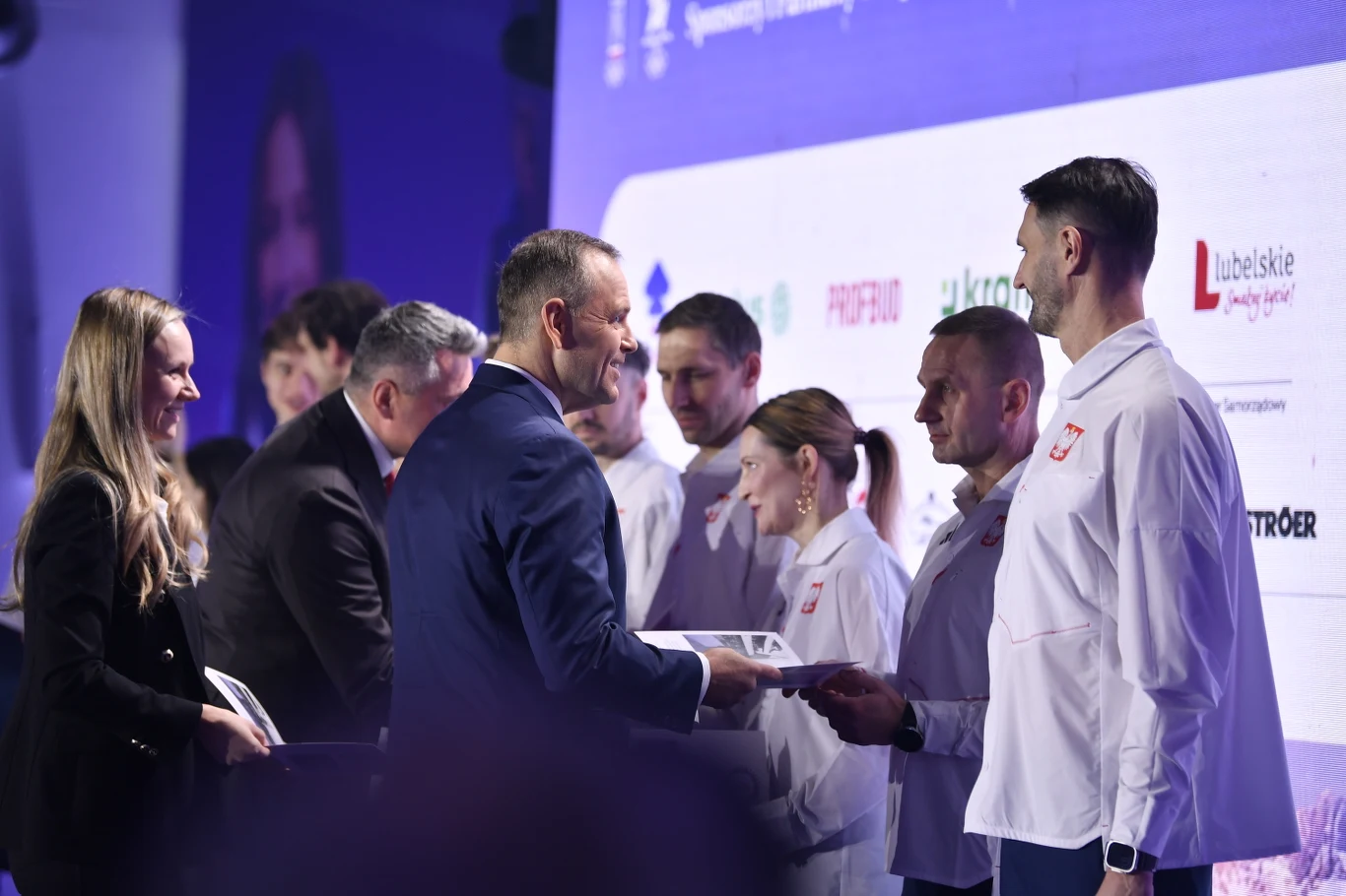 Prezydent Karol Nawrocki na ślubowaniu olimpijczyków Grupa osób ubranych w oficjalne stroje sportowe otrzymuje nagrody lub dyplomy od przedstawicieli podczas oficjalnej uroczystości na tle tablicy z logotypami partnerów i sponsorów.