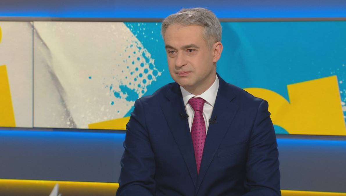 Mężczyzna w granatowym garniturze i r&oacute;żowym krawacie siedzi w studiu telewizyjnym.