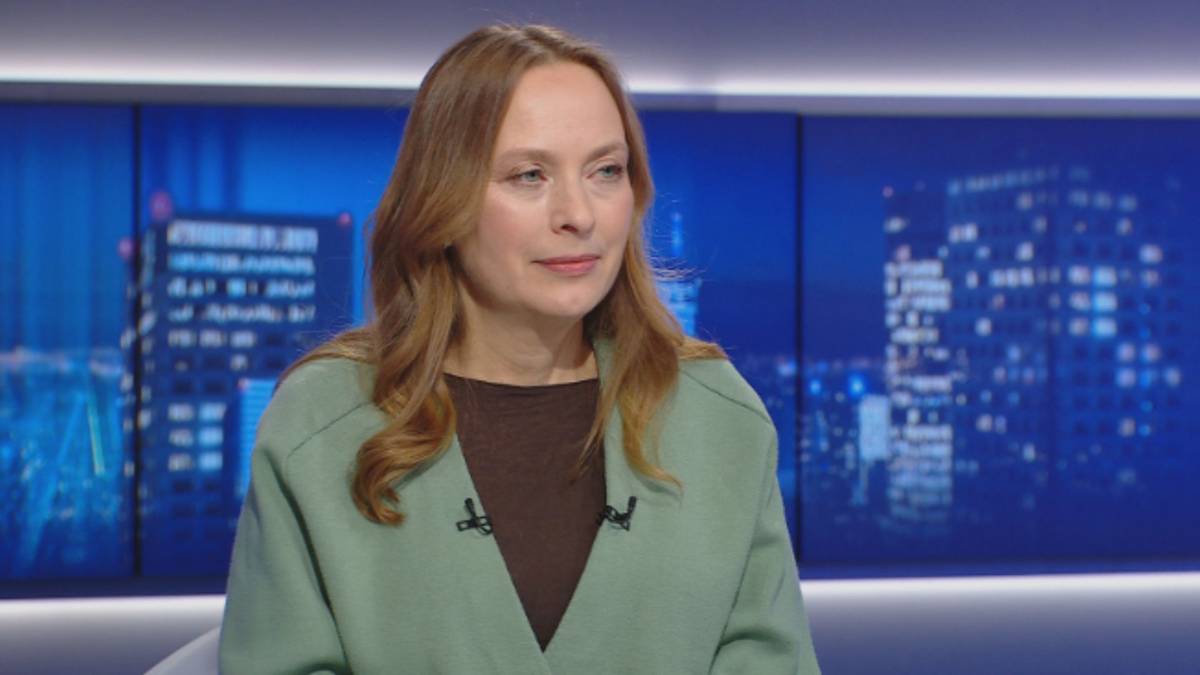 Kobieta udzielająca wywiadu w studiu telewizyjnym, siedzi na krześle, w tle widoczne są ekrany z miejskim krajobrazem.