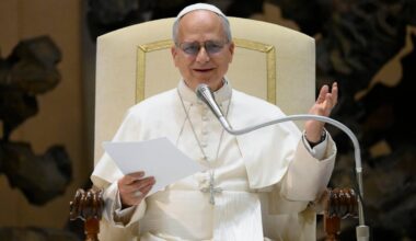 Papież Franciszek w białej szacie siedzi na tronie, czyta z kartki i przemawia do mikrofonu.