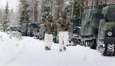 Dw&oacute;ch żołnierzy w zimowym kamuflażu, z karabinami, idzie przez zaśnieżony las w kierunku rzędu pojazd&oacute;w gąsienicowych.