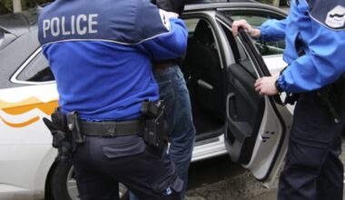 Dw&oacute;ch policjant&oacute;w w niebieskich kurtkach wyprowadza mężczyznę z radiowozu.