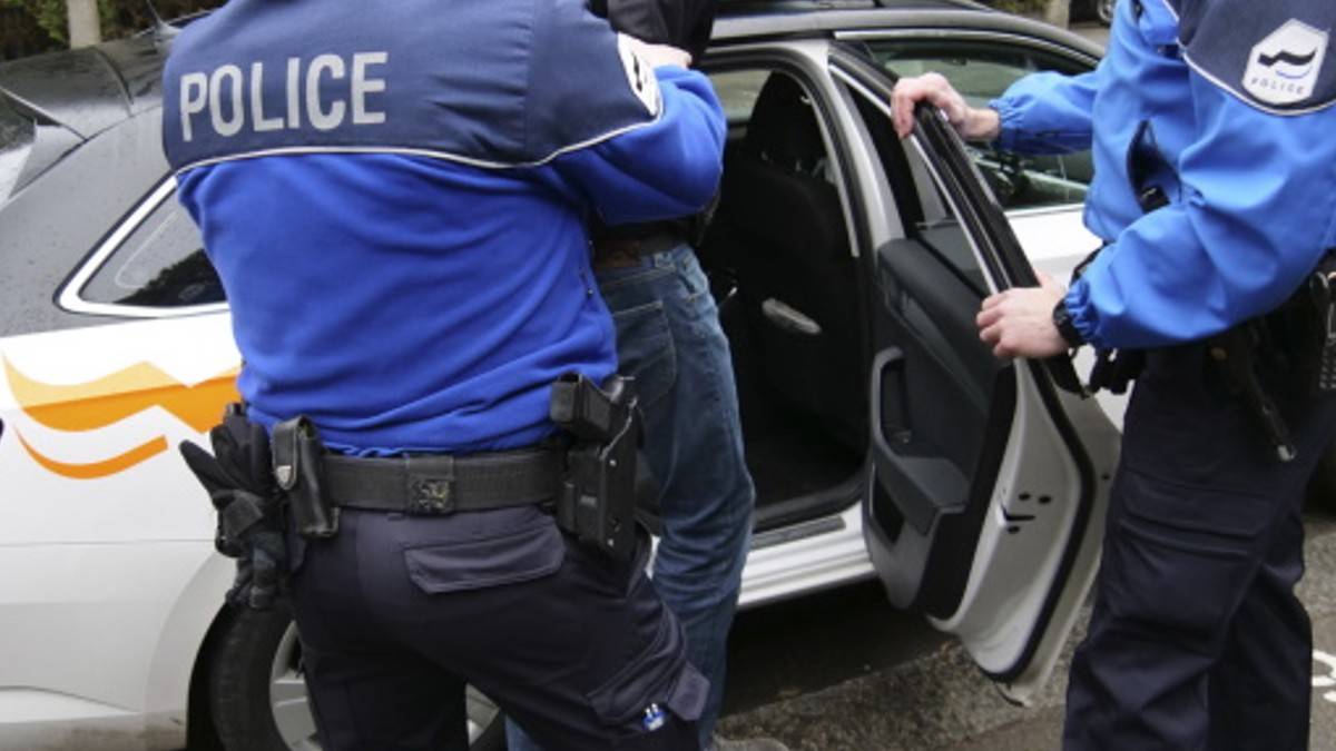 Dw&oacute;ch policjant&oacute;w w niebieskich kurtkach wyprowadza mężczyznę z radiowozu.