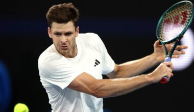 Gracz tenisa w białej koszulce wykonuje uderzenie rakietą, z piłką w powietrzu.