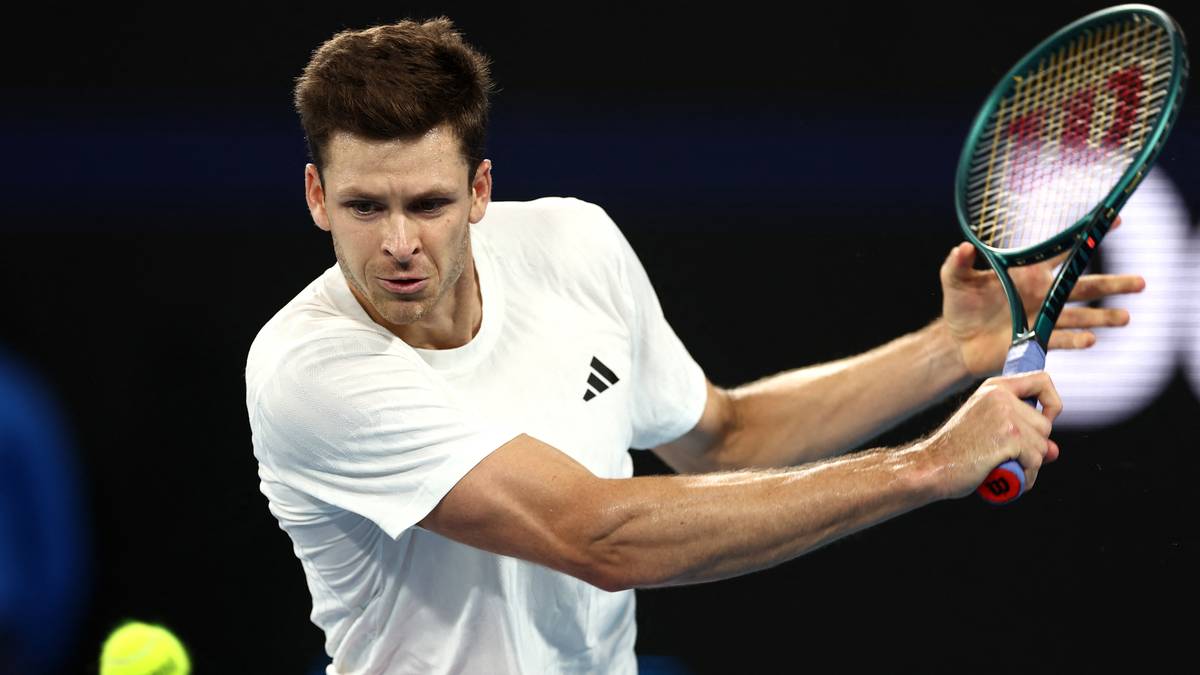 Gracz tenisa w białej koszulce wykonuje uderzenie rakietą, z piłką w powietrzu.