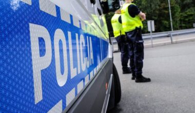 Fragment radiowozu policyjnego z napisem "POLICJA" i stojący obok policjanci w kamizelkach odblaskowych.