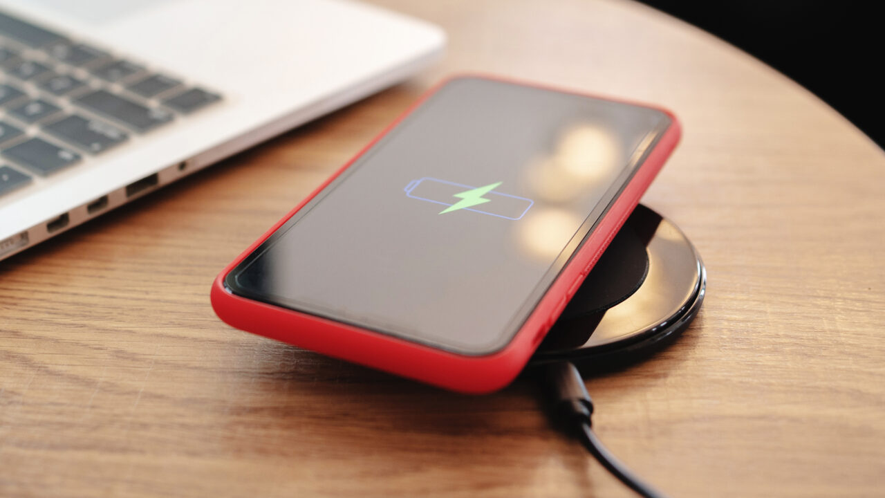 Smartfon w czerwonym etui ładowany bezprzewodowo na czarnej podkładce, na ekranie ikona ładowania, obok laptop.