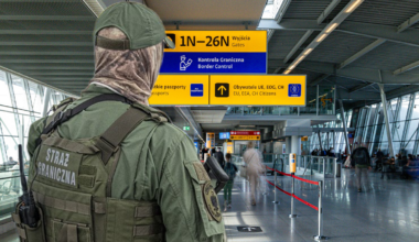 Funkcjonariusz Straży Granicznej w mundurze taktycznym z kamizelką i czapką, stoi na lotnisku. Za nim znajdują się tablice informacyjne z napisami "Kontrola Graniczna" i "Bramki".