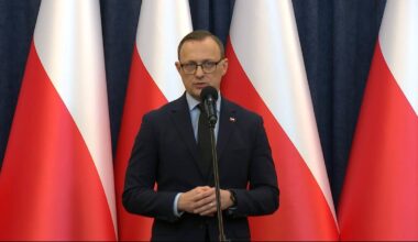 Mężczyzna w garniturze i okularach stoi przed mikrofonem, przemawia na tle flag Polski.