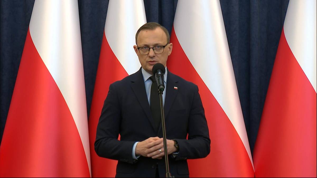 Mężczyzna w garniturze i okularach stoi przed mikrofonem, przemawia na tle flag Polski.