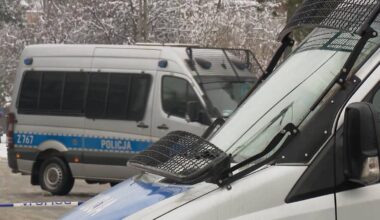 Dwa białe furgony policyjne zaparkowane na zewnątrz, jeden z nich jest w pełni widoczny z przodu, drugi z tyłu, oba mają logo policji.