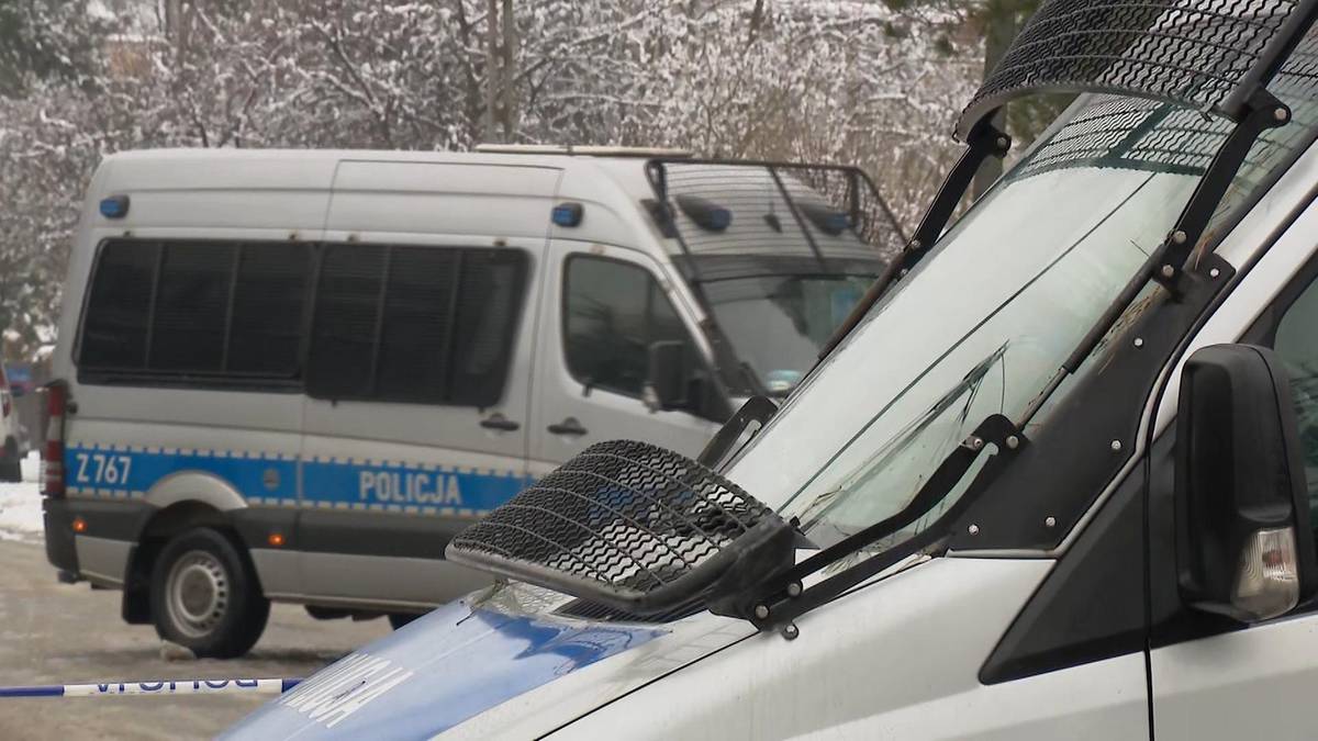 Dwa białe furgony policyjne zaparkowane na zewnątrz, jeden z nich jest w pełni widoczny z przodu, drugi z tyłu, oba mają logo policji.