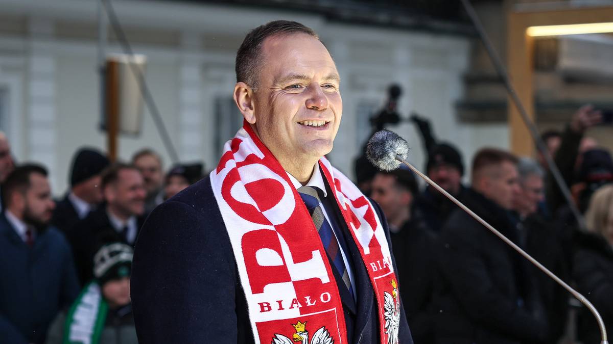 Prezydent Karol Nawrocki w szaliku biało-czerwonym z godłem Polski przemawia do zgromadzonego tłumu.