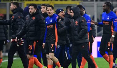 Grupa piłkarzy w czarnych kurtkach z niebieskimi i pomarańczowymi detalami, idąca razem na boisku.