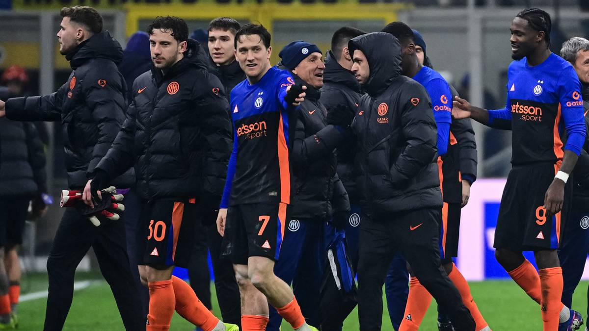 Grupa piłkarzy w czarnych kurtkach z niebieskimi i pomarańczowymi detalami, idąca razem na boisku.