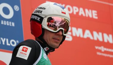 Skoczek narciarski w kasku i goglach, w tle czerwony baner z napisami.