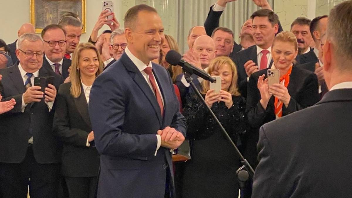 Grupa polityk&oacute;w w garniturach i eleganckich strojach stoi w pomieszczeniu, niekt&oacute;rzy patrzą w kierunku m&oacute;wcy. Jeden z mężczyzn w środku, w garniturze, przemawia do mikrofonu.