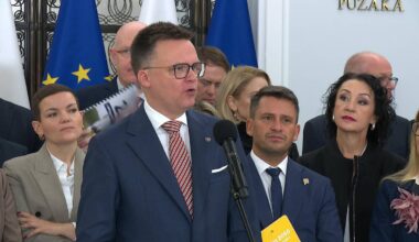 Mężczyzna w okularach i garniturze przemawia do mikrofonu, otoczony przez innych ludzi.