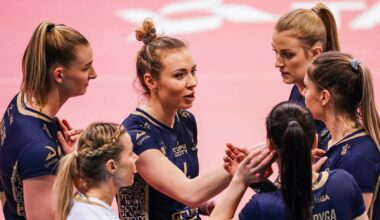 Grupa siatkarek w ciemnogranatowych strojach sportowych dyskutuje w k&oacute;łku na boisku.