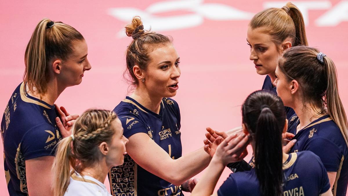 Grupa siatkarek w ciemnogranatowych strojach sportowych dyskutuje w k&oacute;łku na boisku.