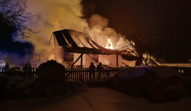 Płonący drewniany budynek w nocy, z ogniem i dymem wydobywającym się z dachu.