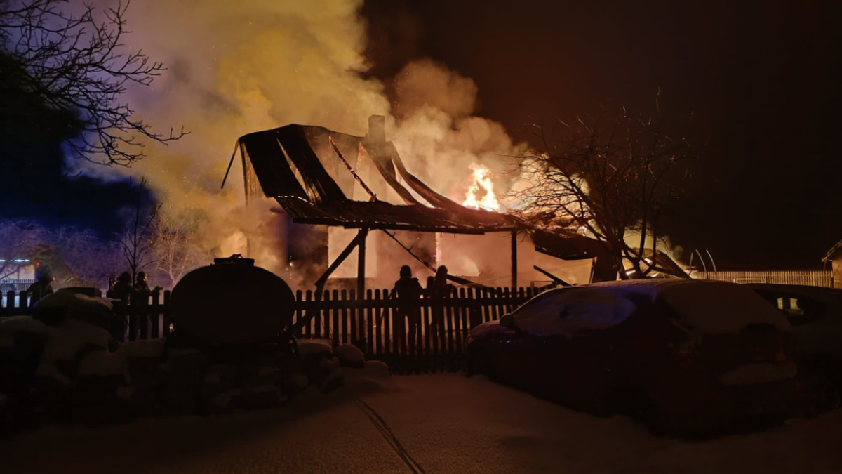 Płonący drewniany budynek w nocy, z ogniem i dymem wydobywającym się z dachu.