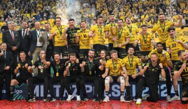 Drużyna siatkarska w ż&oacute;łtych strojach z medalami na szyjach świętuje zwycięstwo, stojąc przed grupą kibic&oacute;w na trybunach.