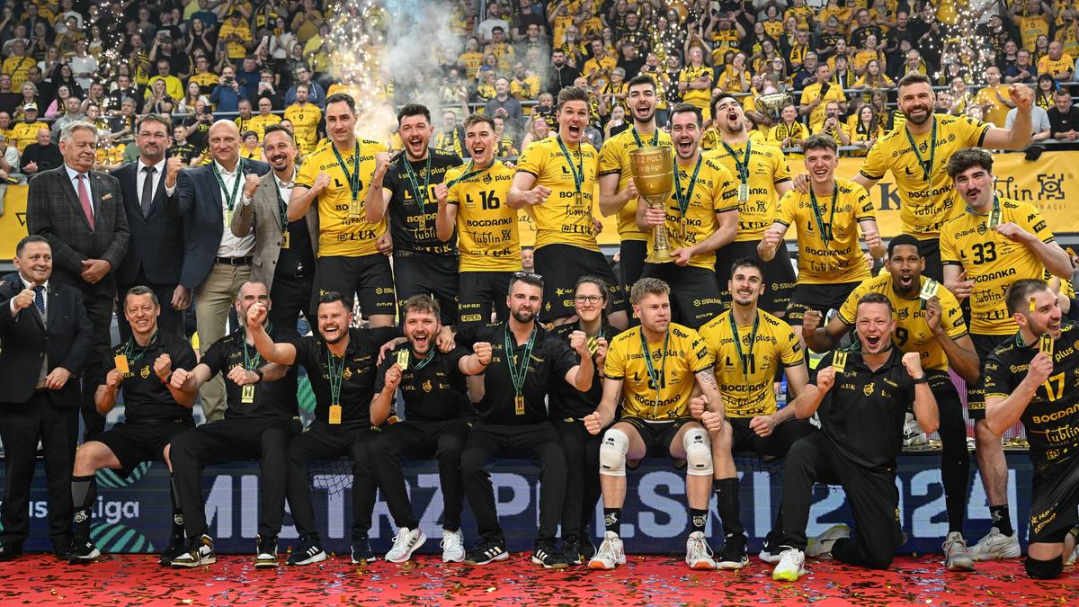 Drużyna siatkarska w ż&oacute;łtych strojach z medalami na szyjach świętuje zwycięstwo, stojąc przed grupą kibic&oacute;w na trybunach.