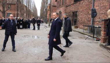 Prezydent Andrzej Duda w ciemnym garniturze i płaszczu, idący w kierunku widza, na tle grupy ludzi w czarnych płaszczach i budynk&oacute;w z cegły.