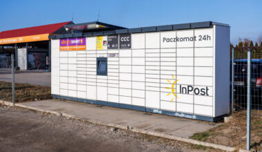 Biały paczkomat InPost 24h z żółtym logo słońca i ekranem obsługi ustawiony na chodniku przy ogrodzeniu i parkingu.