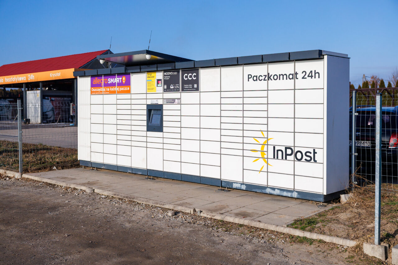 Biały paczkomat InPost 24h z żółtym logo słońca i ekranem obsługi ustawiony na chodniku przy ogrodzeniu i parkingu.