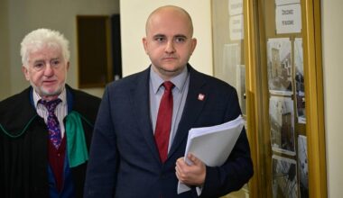 Poseł PiS Dariusz Matecki w garniturze i czerwonym krawacie trzyma dokumenty, obok stoi mężczyzna w todze prawniczej.