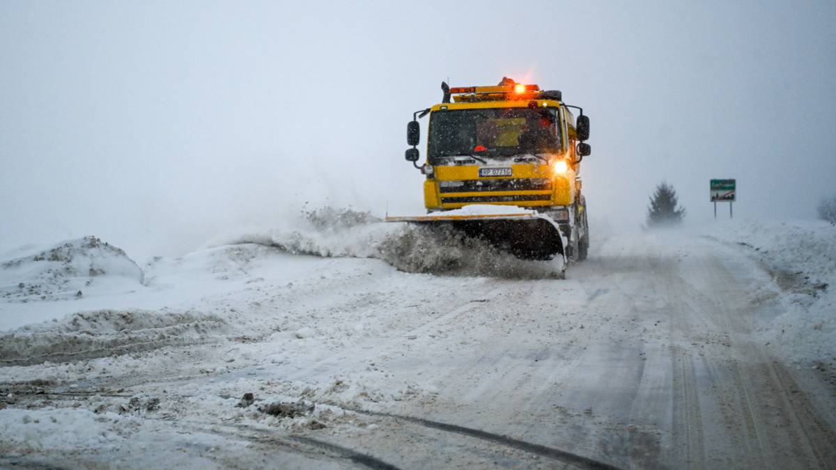 Ż&oacute;łty pług śnieżny z włączonymi światłami ostrzegawczymi usuwa śnieg z zaśnieżonej drogi.