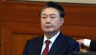 Portret mężczyzny w ciemnym garniturze i czerwonym krawacie, siedzącego w pomieszczeniu o drewnianych panelach.