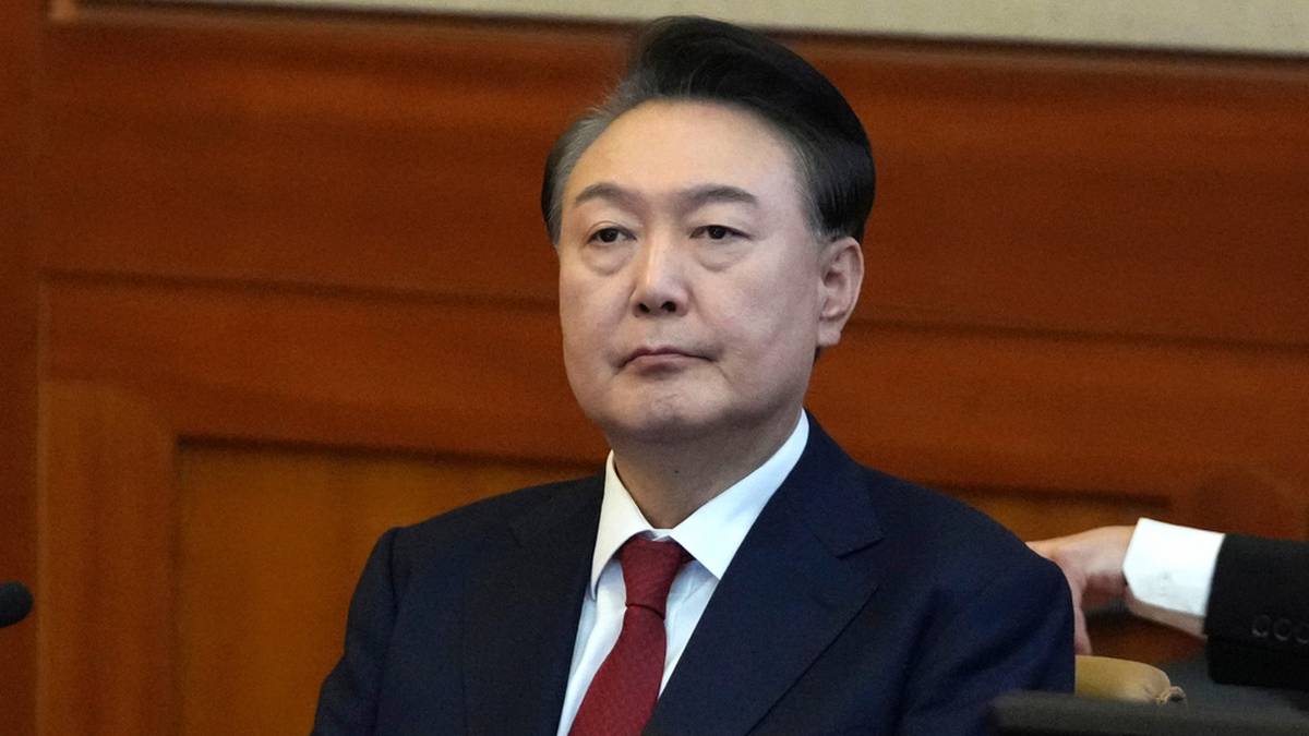 Portret mężczyzny w ciemnym garniturze i czerwonym krawacie, siedzącego w pomieszczeniu o drewnianych panelach.