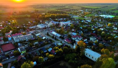 Widok z lotu ptaka na miasto o zachodzie słońca, z widocznymi budynkami, drogami i otaczającą zielenią.