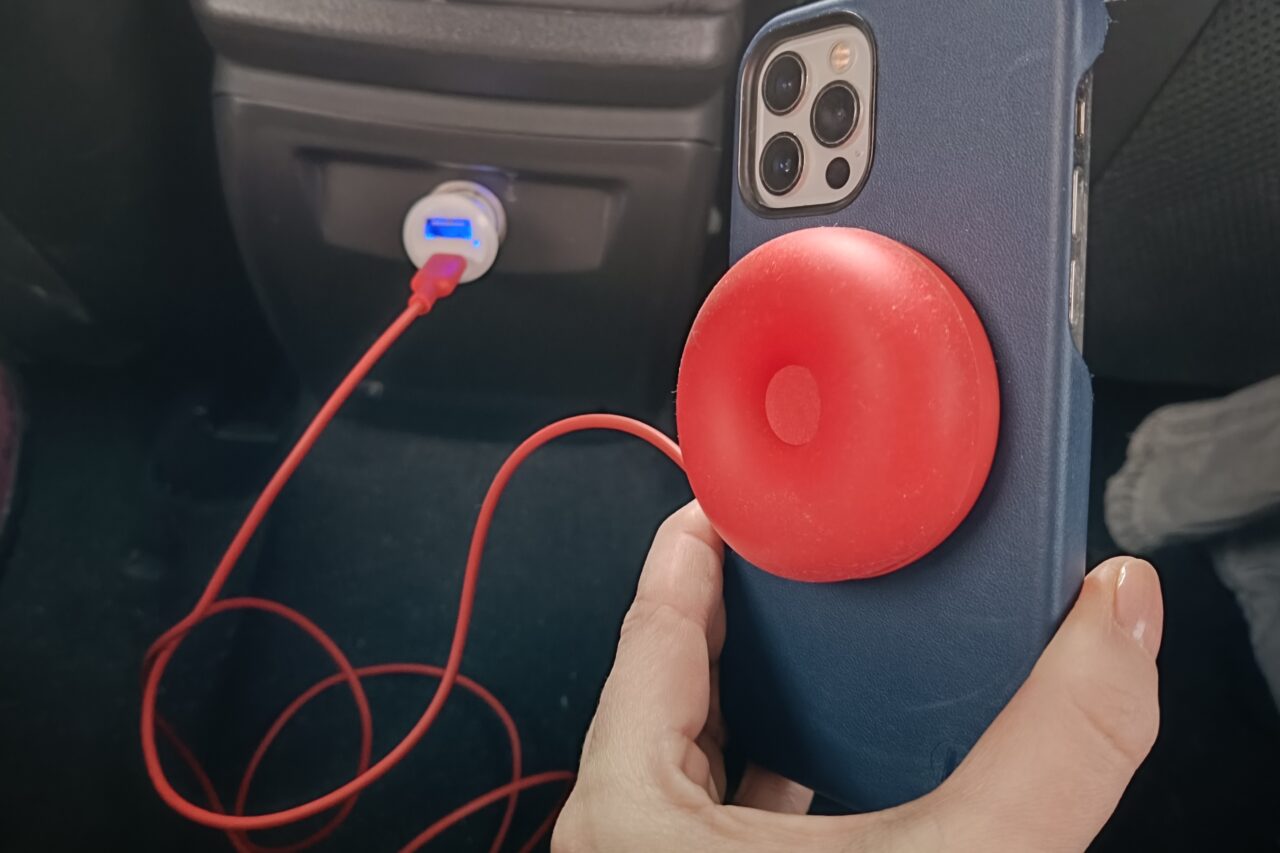 Ręka trzyma smartfon w granatowym etui z czerwonym okrągłym ładowaczem podłączonym kablem do ładowarki samochodowej.