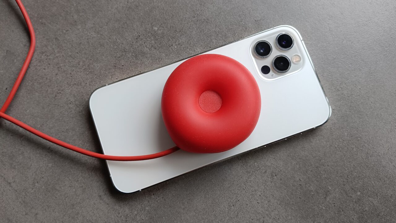 Biały smartfon z potrójnym aparatem na szarej powierzchni z przyczepioną czerwoną, pączkowatą ładowarką z kablem.