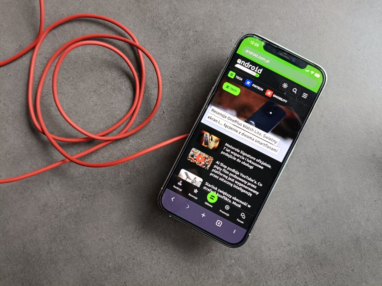 Smartfon z otwartą stroną android leżący ukośnie na szarym blacie obok luźno zwiniętego czerwonego kabla.