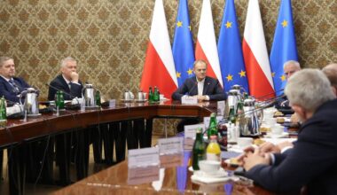 Grupa mężczyzn siedzi przy stole podczas spotkania, za nimi widoczne są flagi Polski i Unii Europejskiej.