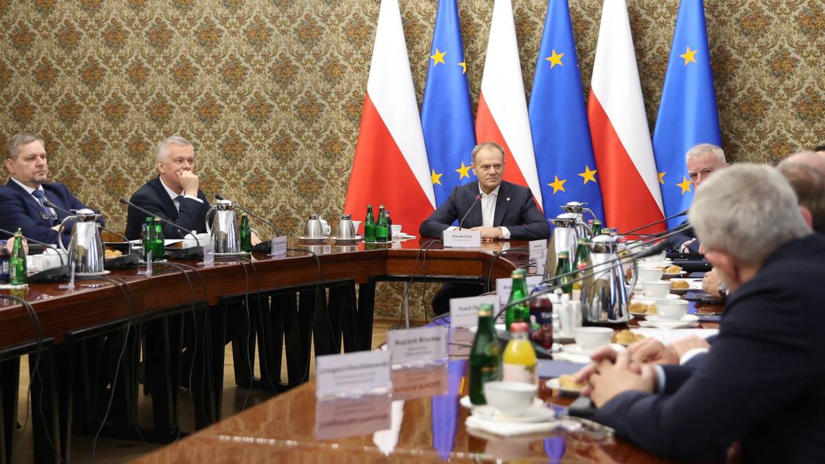 Grupa mężczyzn siedzi przy stole podczas spotkania, za nimi widoczne są flagi Polski i Unii Europejskiej.