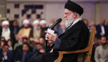 Starszy mężczyzna w czarnym nakryciu głowy i szacie przemawia do mikrofonu.