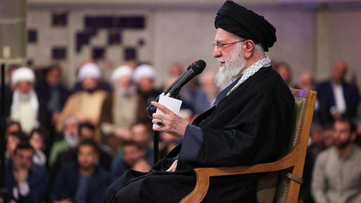 Starszy mężczyzna w czarnym nakryciu głowy i szacie przemawia do mikrofonu.