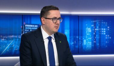 Mężczyzna w okularach i garniturze siedzi przed niebieskim tłem.