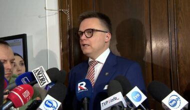 Mężczyzna w okularach i garniturze w otoczeniu mikrofon&oacute;w medi&oacute;w.