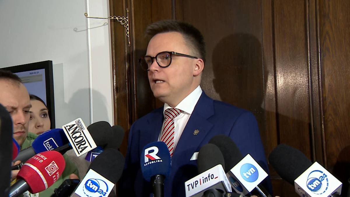 Mężczyzna w okularach i garniturze w otoczeniu mikrofon&oacute;w medi&oacute;w.